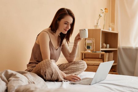 Foto de Acogedor retrato de larga duración de la mujer joven usando el ordenador portátil en la cama en el extremo de casa disfrutando de café, espacio para copiar - Imagen libre de derechos