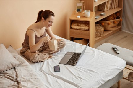 Retrato de alto ángulo de una joven sonriente comiendo fideos para llevar y viendo películas a través de un ordenador portátil en casa, espacio para copiar