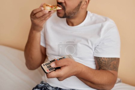 Téléchargez les photos : Gros plan d'un homme tatoué mangeant de la pizza à la maison et tenant la télécommande tout en appréciant le week-end de détente - en image libre de droit