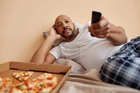 Portrait of bored adult man watching TV at home and lying on bed with pizza, copy space