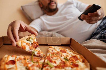 Téléchargez les photos : Gros plan de l'homme adulte prenant tranche de pizza tout en appréciant week-end paresseux à la maison et regarder la télévision, copier l'espace - en image libre de droit