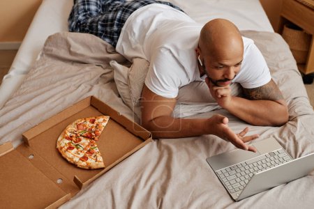 Foto de Vista superior del hombre negro adulto acostado en la cama en casa y disfrutando de un fin de semana perezoso con pizza - Imagen libre de derechos