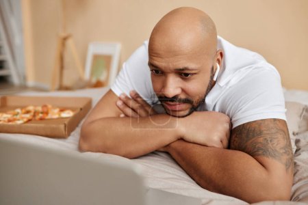 Foto de Primer plano del hombre negro adulto viendo videos a través de la computadora portátil acostado en la cama en casa durante el fin de semana perezoso - Imagen libre de derechos