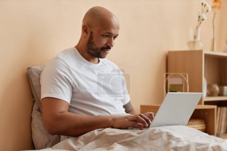 Side view portrait of bald black man using laptop in bed in cozy home, copy space