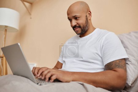 Portrait of smiling black man using laptop in bed in cozy home, copy space