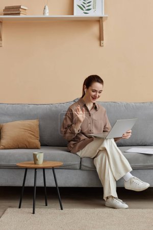 Photo for Vertical full length portrait of young elegant businesswoman enjoying work from home and sitting on comfortable sofa waving to online meeting - Royalty Free Image