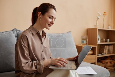 Photo for Side view portrait of young elegant businesswoman enjoying work from home and sitting on comfortable sofa with laptop speaking in online meeting - Royalty Free Image