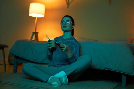 Foto de Retrato de larga duración de una mujer joven viendo la televisión en la oscuridad y comiendo fideos para llevar mientras está sentada en el suelo en casa, espacio para copiar - Imagen libre de derechos