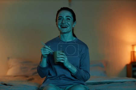 Front view portrait of smiling adult woman watching movies at home in dark and eating takeout food