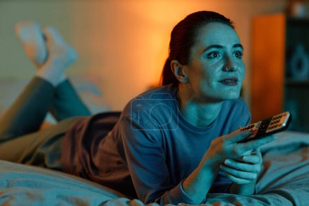 Téléchargez les photos : Portrait de femme adulte allongée sur le lit à la maison et regardant la télévision dans l'obscurité - en image libre de droit
