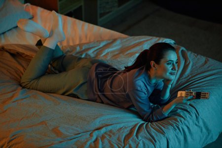 Téléchargez les photos : Portrait grand angle de femme adulte regardant la télévision dans l'obscurité tout en étant couché sur le lit à la maison, espace de copie - en image libre de droit