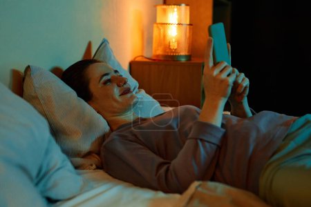 Foto de Retrato de vista lateral de una joven sonriente usando un teléfono inteligente en la cama por la noche - Imagen libre de derechos