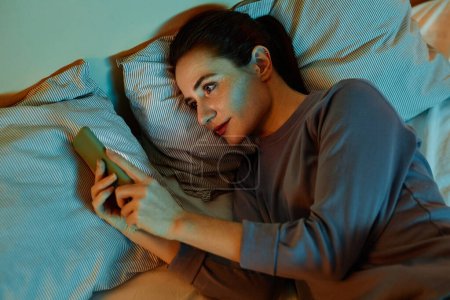 Photo for Top view portrait of smiling adult woman using smartphone in bed at night reading text messages and scrolling social media - Royalty Free Image