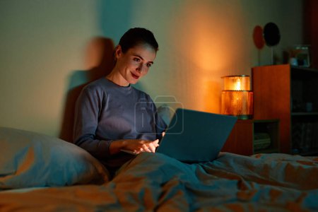 Téléchargez les photos : Portrait de femme adulte souriante utilisant un ordinateur portable au lit la nuit, espace de copie - en image libre de droit