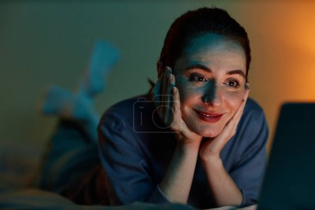 Téléchargez les photos : Gros plan portrait de jeune femme binge regarder des films la nuit tandis que couché sur le lit avec ordinateur portable - en image libre de droit