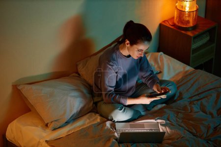Foto de Retrato de ángulo alto de una mujer joven que trabaja hasta tarde en casa mientras está sentada en la cama con computadora portátil y teléfono inteligente - Imagen libre de derechos