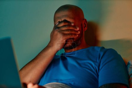 Foto de Retrato del hombre negro adulto usando el ordenador portátil en la cama por la noche y frotando los ojos - Imagen libre de derechos