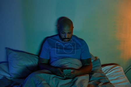 Foto de Retrato de hombre adulto negro usando teléfono inteligente en la cama por la noche que sufre de insomnio - Imagen libre de derechos