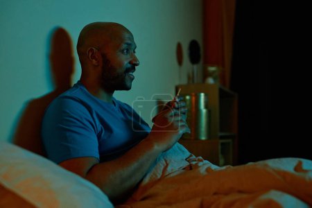 Téléchargez les photos : Portrait latéral d'un homme noir souriant regardant la télévision au lit la nuit, restant debout tard, copiant l'espace - en image libre de droit