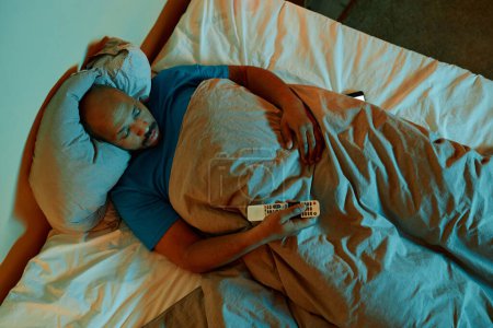 Téléchargez les photos : Vue du dessus à l'homme noir regardant la télévision au lit la nuit souffrant d'insomnie - en image libre de droit