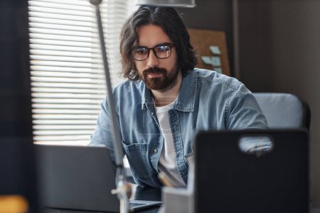 Téléchargez les photos : Portrait d'un homme barbu portant des lunettes à l'aide d'ordinateurs au bureau tout en travaillant sur un projet informatique - en image libre de droit