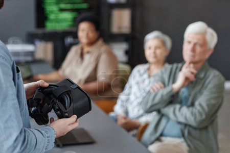 Closeup of man holding VR headset presenting virtual reality technology to group of senior people, copy space