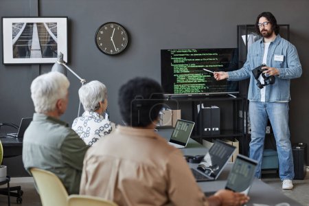Téléchargez les photos : Portrait complet de l'homme barbu pointant vers l'écran avec le code tout en enseignant aux personnes âgées dans la classe de programmation informatique, espace de copie - en image libre de droit