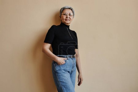 Téléchargez les photos : Portrait taille haute de la femme âgée asiatique moderne regardant la caméra avec confiance debout contre le mur - en image libre de droit
