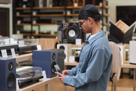 Photo for Side view portrait of young black man looking at vinyl record player in music store and taking picture of price tag with smartphone - Royalty Free Image
