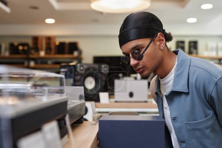 Téléchargez les photos : Portrait latéral de jeune homme noir regardant des lecteurs de disques et du matériel dans un magasin de musique, espace de copie - en image libre de droit
