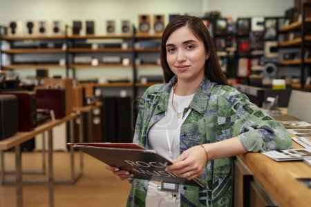 Waist up portrait of young woman working in music store amd holding record smiling at camera, copy space