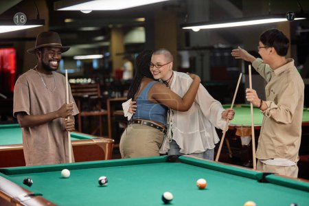 Photo for Portrait of two young women embracing at pool table enjoying game of billiards with diverse group of friends - Royalty Free Image