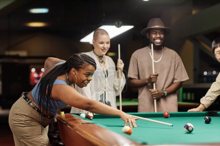 Foto de Retrato de vista lateral de una mujer afroamericana sonriente jugando al billar en la mesa en el club nocturno con un diverso grupo de amigos copiar espacio - Imagen libre de derechos