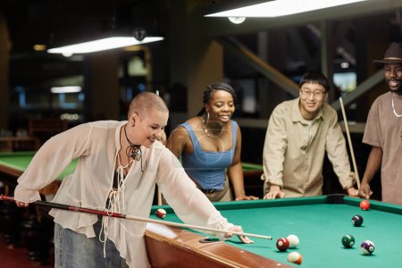 Photo for Side view portrait of smiling young bald woman playing pool at table in nightclub with diverse group of friends copy space - Royalty Free Image