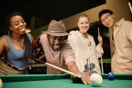 Photo for Multiethnic group of friends playing pool together with focus on smiling Black man hitting ball with cue stick copy space - Royalty Free Image