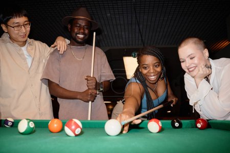 Multiethnische Gruppe lächelnder junger Leute spielt Pool zusammen mit schwarzer Frau, die Ball mit Queue-Stick schlägt