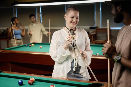 Photo for Waist up portrait of bald young woman chatting with friend and smiling while enjoying game of pool together in low light copy space - Royalty Free Image