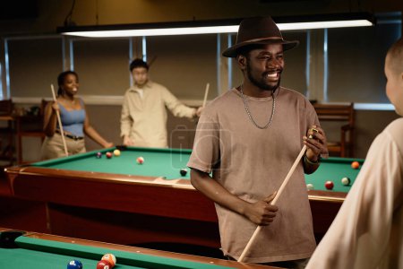 Foto de Cintura hacia arriba retrato de hombre adulto afroamericano charlando con un amigo y sonriendo mientras disfrutan del juego de billar juntos en espacio de copia de poca luz - Imagen libre de derechos