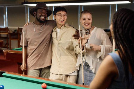 Téléchargez les photos : Portrait taille haute d'un groupe multiethnique d'amis bavardant et souriant tout en appréciant le jeu de billard ensemble dans une faible lumière - en image libre de droit
