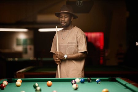Photo for Waist up portrait of adult African American man wearing hat and holding cue stick while standing by table in pool club copy space - Royalty Free Image