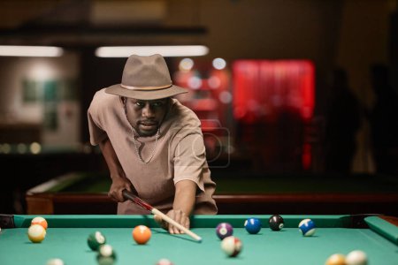 Photo for Waist up portrait of adult African American man wearing hat and hitting ball with cue stick while playing pool copy space - Royalty Free Image