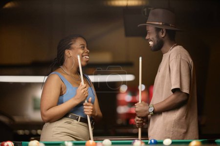 Photo for Side view portrait of smiling African American couple chatting by pool table in nightclub - Royalty Free Image
