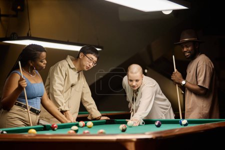 Foto de Retrato de grupo multiétnico de amigos jugando al billar juntos por mesa en discoteca en tonos apagados - Imagen libre de derechos