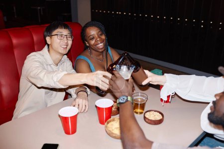 Foto de Grupo multiétnico de amigos sacudiendo botellas de cerveza y animando sentados en la mesa del comedor juntos - Imagen libre de derechos
