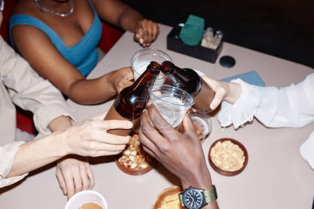 Téléchargez les photos : Gros plan d'amis multiethniques accrochant des bouteilles de bière et acclamant sur la table à manger ensemble - en image libre de droit