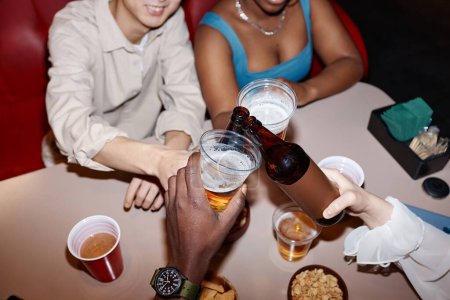 Foto de Primer plano de un grupo diverso de jóvenes sosteniendo botellas de cerveza y animando a la mesa de comedor juntos. - Imagen libre de derechos