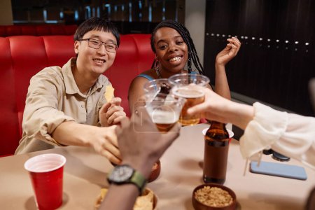 Foto de Grupo multiétnico de jóvenes sacudiendo botellas de cerveza y animando sentado en la mesa del comedor - Imagen libre de derechos