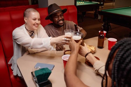 Foto de Grupo multiétnico de amigos tintineando vasos de cerveza y animando sentado en la mesa del comedor - Imagen libre de derechos