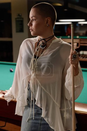 Vertical side view portrait of confident young woman with buzz cut holding cue stick in pool club