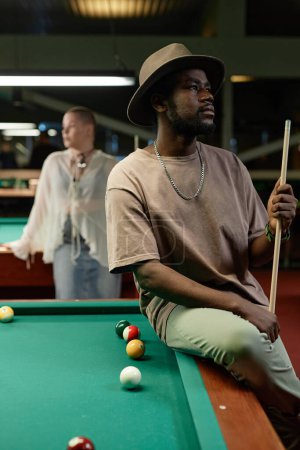 Téléchargez les photos : Portrait latéral d'un homme noir adulte portant un chapeau posant sur une table de billard dans un bar - en image libre de droit